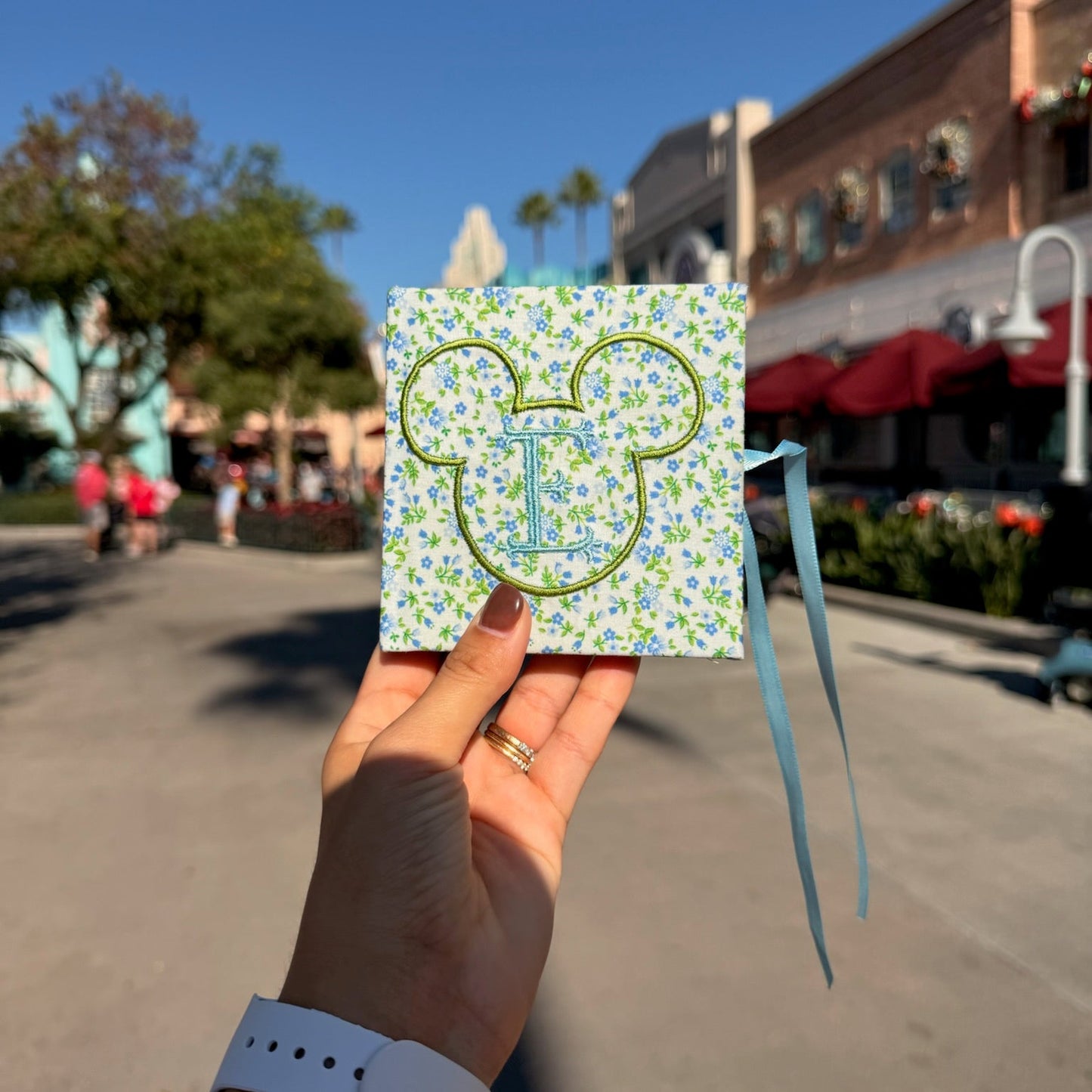 Hand holding a decorative card with a floral pattern and letter 'T' in a park setting